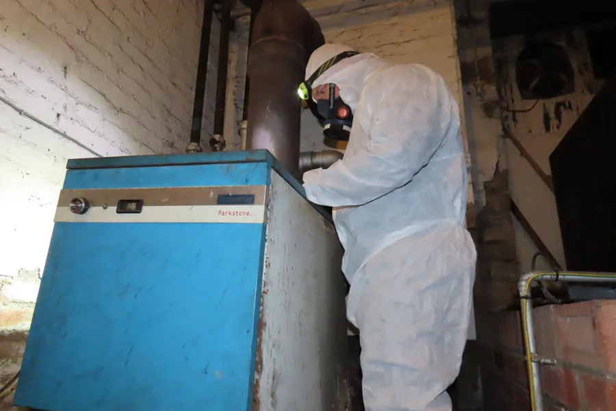 A qualified asbestos surveyor wearing protective coveralls and respiratory equipment inspects an old boiler for potential asbestos-containing materials.