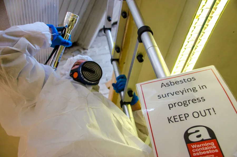 A qualified asbestos surveyor wearing protective coveralls and respiratory equipment uses a ladder to inspect a high-level area during an asbestos survey.
