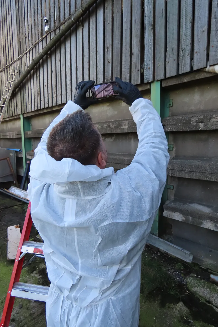 A qualified asbestos surveyor wearing protective coveralls photographs the exterior materials of an agricultural building during an asbestos survey.