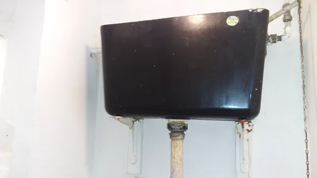 An older black toilet cistern, likely manufactured from asbestos-containing reinforced plastic, with associated pipework and pull-chain flushing mechanism. The cistern should not be drilled, cut or damaged without appropriate asbestos precautions.