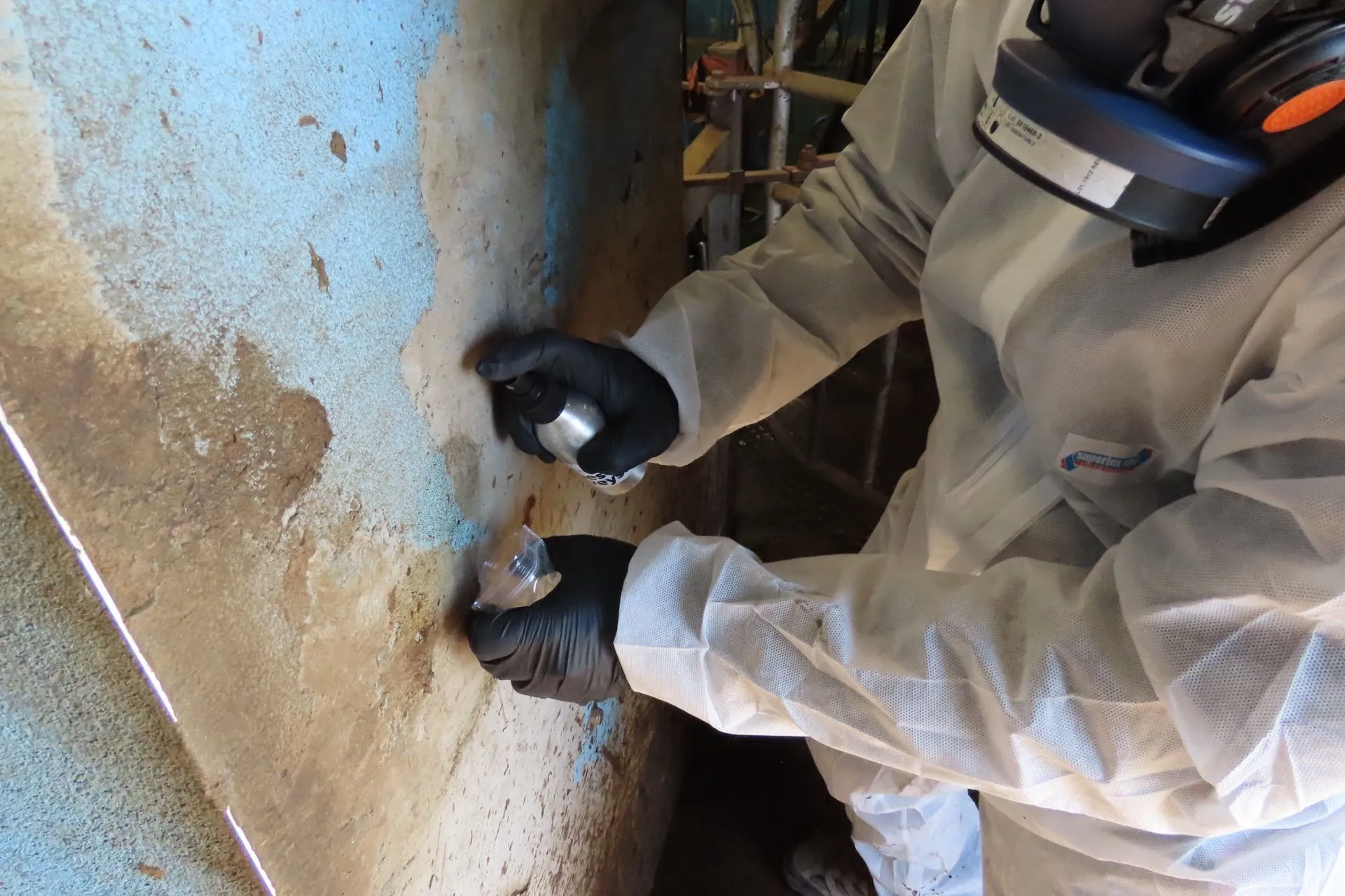 Surveyor in coveralls and a respirator damping down suspect material before taking a sample into a sealed bag