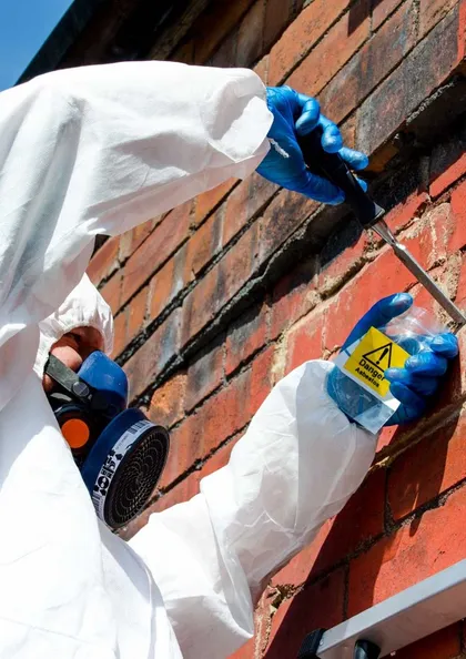 Analyst in a protective coverall, respirator and gloves taking an asbestos sample from an external brick wall