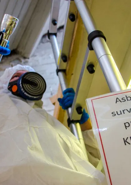 Asbestos surveyor in a protective coverall and respirator working beside an 'Asbestos surveying in progress' warning sign