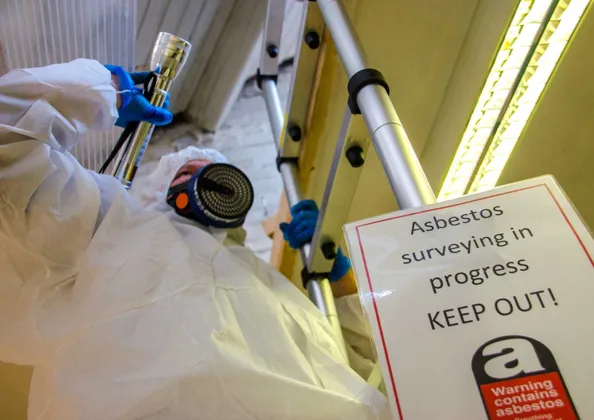 Asbestos surveyor in a protective coverall and respirator working beside an 'Asbestos surveying in progress' warning sign